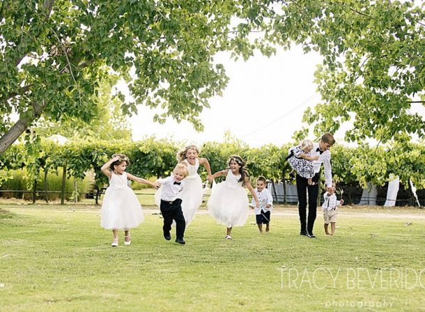 Little flowergirls and pageboys holding hands and running and laughing playfully under trees with a backdrop of grapevines.