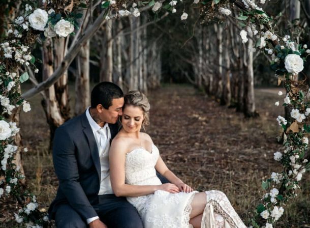 Bride and groom on a circular floral swing at The Vinegrove's Mudgee wedding venue, with the groom intimately whispering in her ear against a serene forest backdrop.