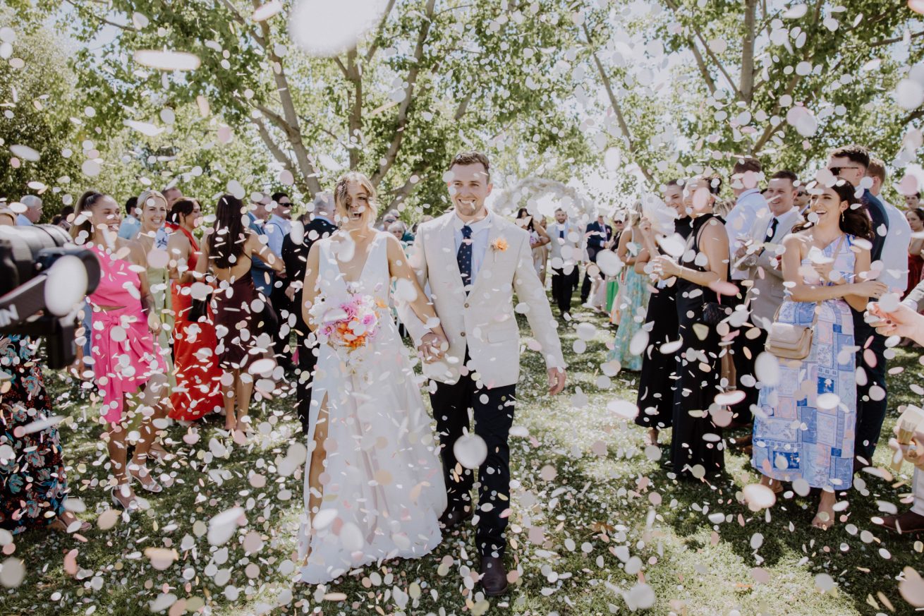 A joyful bride and groom laughing as they walk down the aisle, with guests celebrating by throwing white rose petals that float down like snowflakes, creating a magical moment.