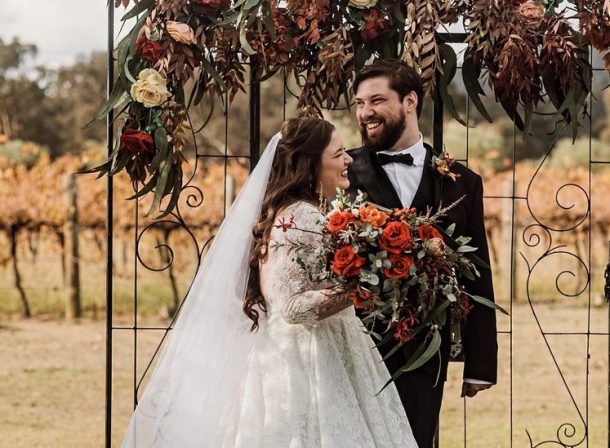 A bride and groom laughing beneath a beautiful autumn floral arrangement adorning a bronzed trellis doorway arch