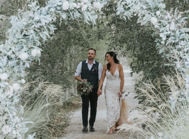 Newlyweds walking hand in hand under a beautifully decorated circular arch adorned with lush greenery and delicate white flowers, surrounded by an avenue of beautiful poplar trees.