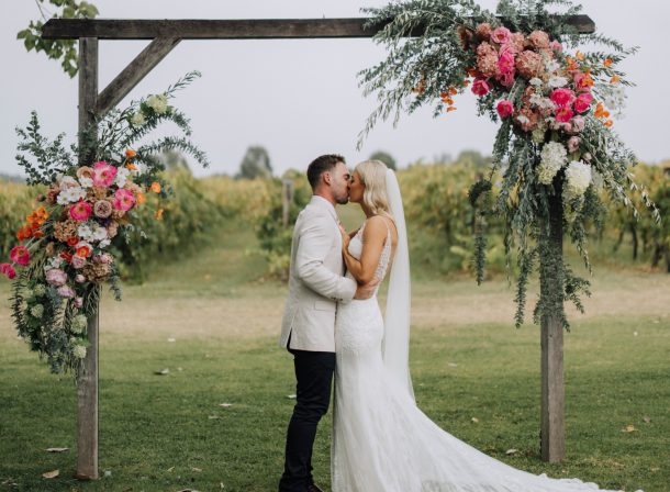 Bride and groom kissing under a beautiful wooden wedding arbor decorated with stunning silk flowers at The Vinegrove wedding venue in Mudgee.