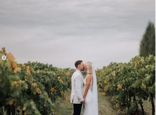 Bride and groom sharing a tender kiss between rows of lush grapevines under a dramatic grey cloudy sky at The Vinegrove Mudgee wedding venue.