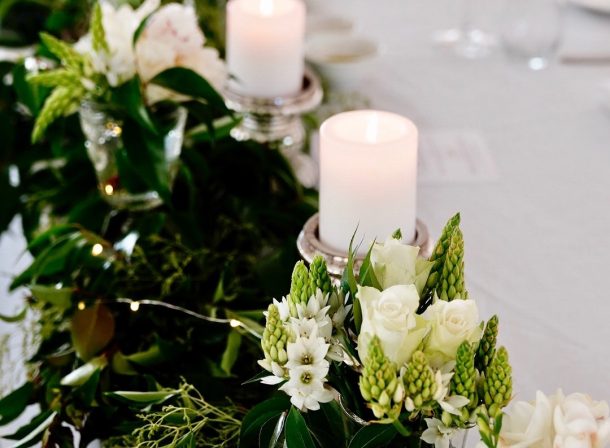 Romantic wedding reception table adorned with cascading greenery and delicate white flowers, embracing softly glowing candles in elegant silver holders.