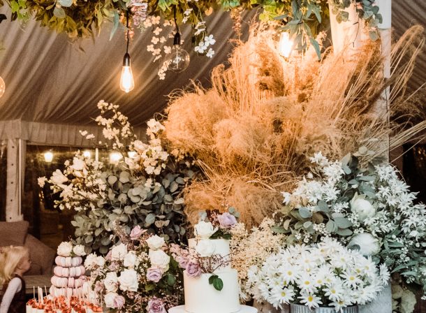 Charming wedding dessert table adorned with a lush floral canopy, featuring a beautiful cake surrounded by elegant roses, delicate white daisies, and rustic pampas grass, all softly illuminated by warm hanging lights.