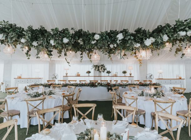 Elegant wedding reception with beautifully decorated tables, featuring lush greenery and white floral arrangements, beneath a stunning canopy of cascading foliage and sparkling chandeliers.