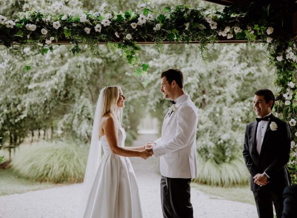 Bride and groom holding hands beneath green and white faux floral wedding arbor, on a rainy wedding day at The Vinegrove's undercover ceremony area.