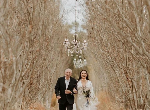 A father walking his daughter down the aisle of a winter poplar avenue adorned with stunning crystal chandeliers hanging above, creating a magical and elegant ambiance.